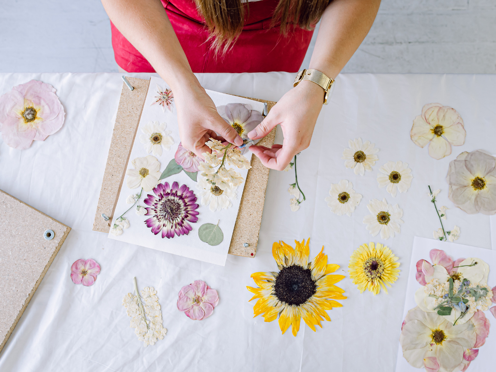 Hands arranging pressed flowers on a work surface beside a flower press